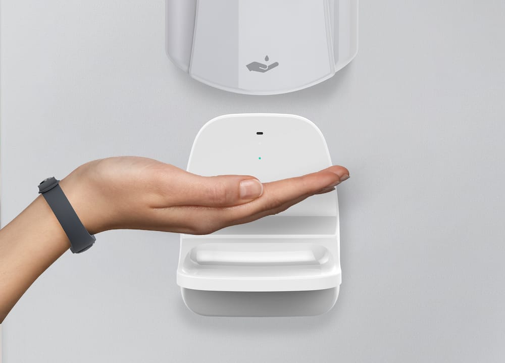 An alcohol solution dispenser with a Sanibit Smart Drip Tray beneath; a hand with a Sanibit Wristband reaches for sanitizer.